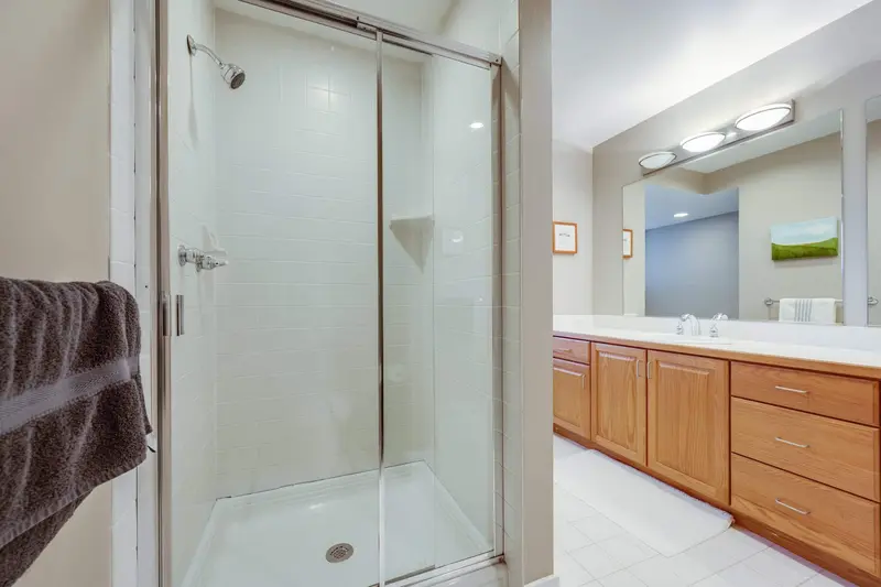 Old glass shower stall beside the end of the light wood vanity and a towel bar