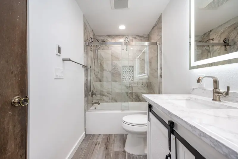View from the doorway over the vanity to the tub with sliding glass and marble-look tile