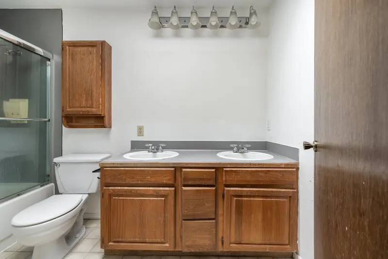Bathroom before the remodel with an oak double vanity, a gray laminate counter and a tub behind framed glass doors