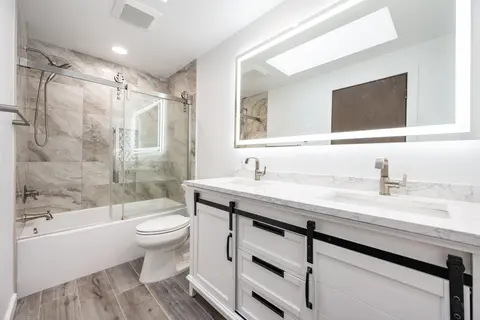 Remodeled bathroom with a white double vanity, a wide LED mirror and a tub behind sliding glass