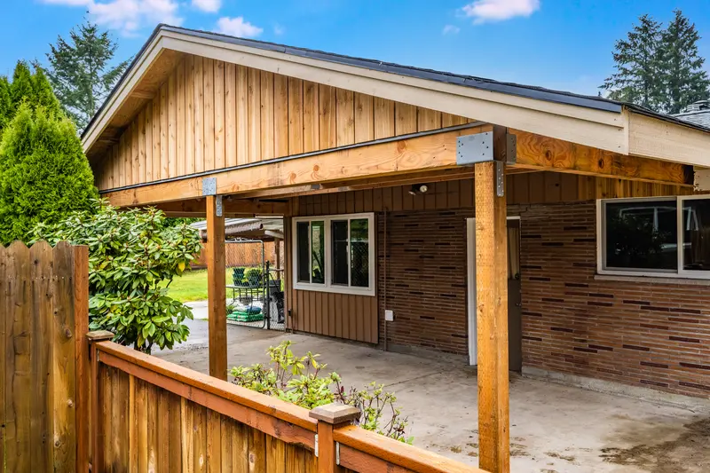 New carport gable end with vertical wood boards and an asphalt shingle roof, on wood posts beside the house