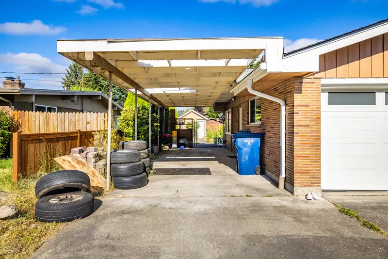 Old flat carport with a white painted frame and translucent roof panels beside a brick garage