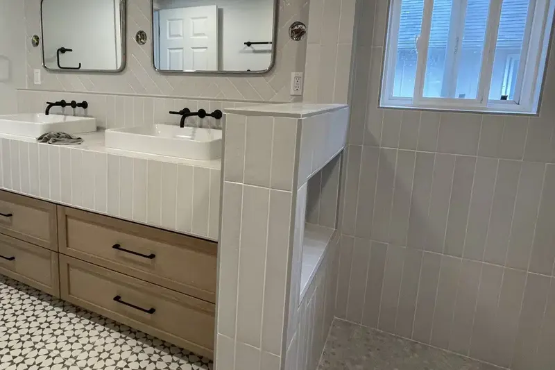 Light oak double vanity with vessel sinks and matte black faucets beside a tiled walk-in shower, from a Seattle bathroom remodel by Volcano Builders