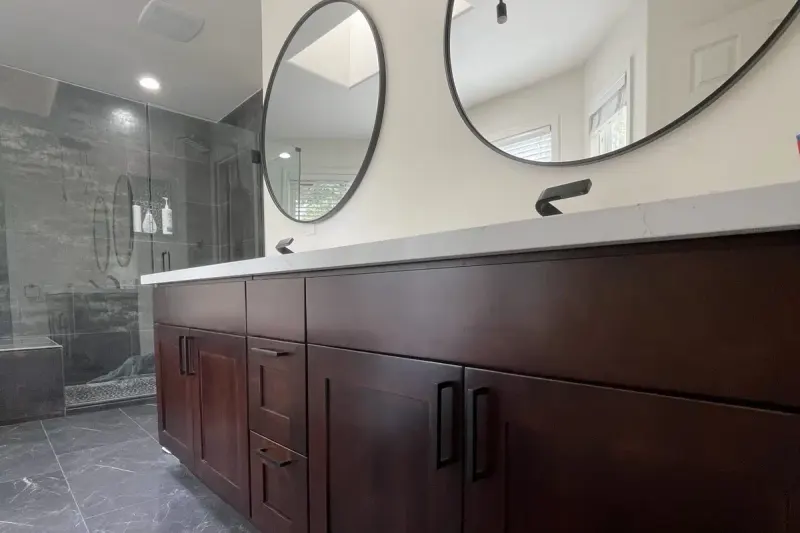 Long dark wood double vanity under two round mirrors, with a glass shower beyond, from an Issaquah bathroom remodel by Volcano Builders