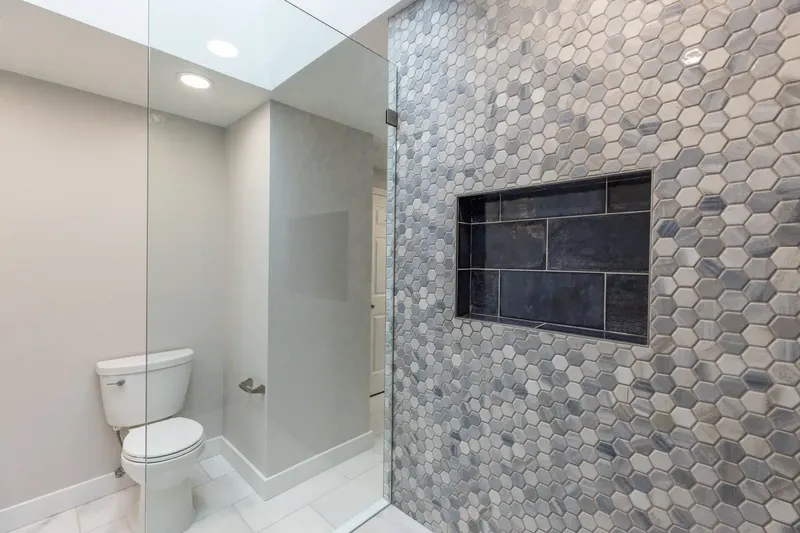 Walk-in shower with a gray hexagon mosaic wall, a dark tile niche and a frameless glass panel, with the toilet alcove and skylight beyond, from a Redmond bathroom remodel by Volcano Builders