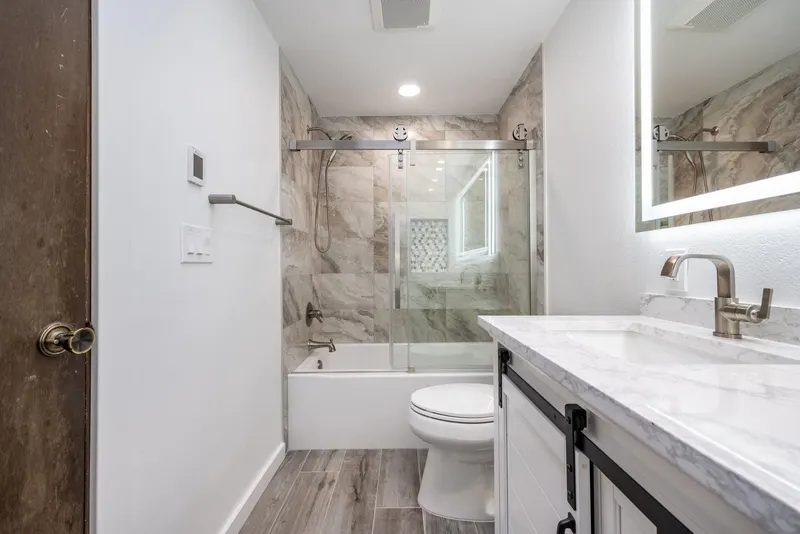 Remodeled bathroom with a white barn door vanity, marble look tub surround behind sliding glass and a lit mirror, from a Tulalip Bay project by Volcano Builders