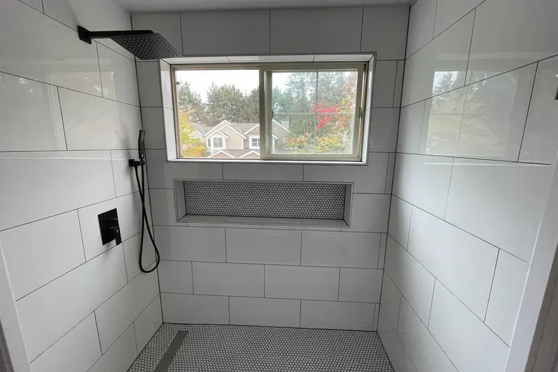 Walk-in shower with large white wall tile, a black rain head, penny tile floor and a window onto trees, from a Redmond remodel by Volcano Builders