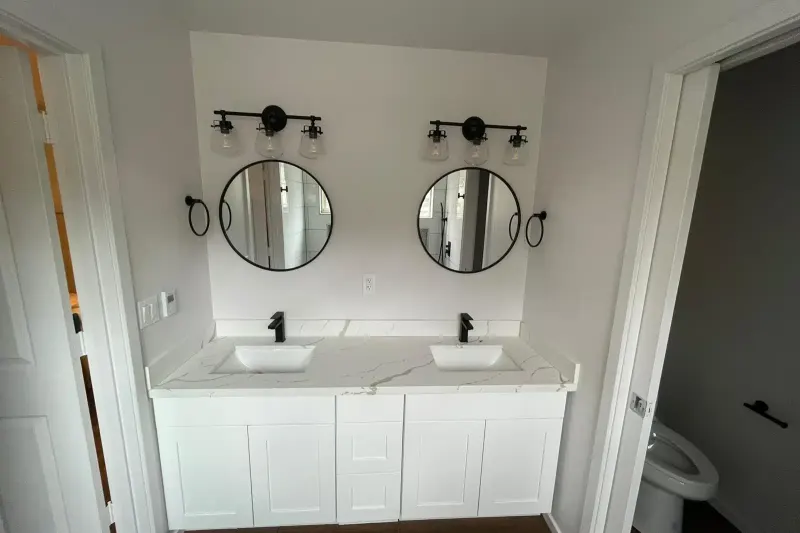Primary bathroom with a white double vanity, quartz top, round black mirrors and matte black fixtures, by Volcano Builders