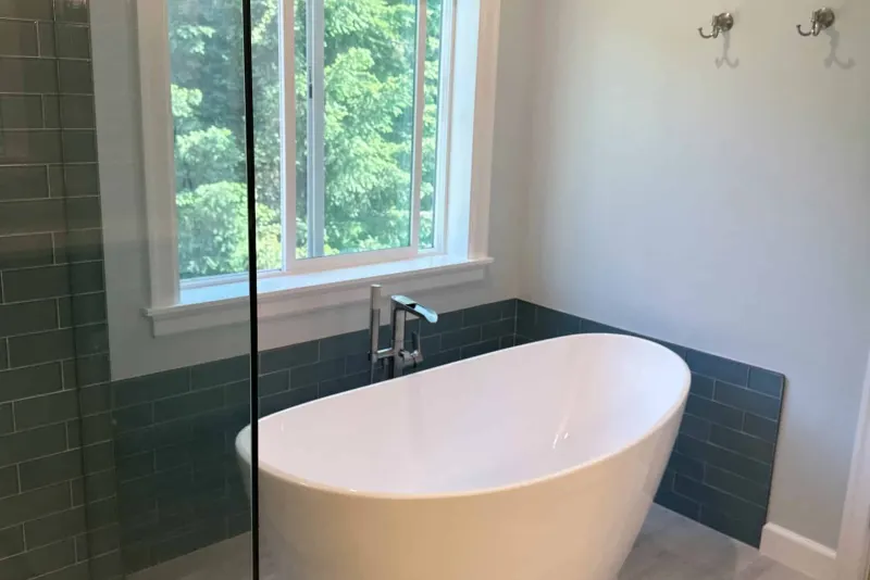Freestanding white soaking tub under a window facing trees, with blue glass tile wainscot, from a Snohomish bathroom remodel by Volcano Builders
