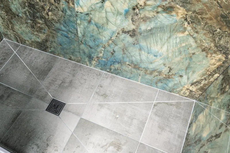 Shower floor in large gray tile with a square black drain against blue-green stone-look walls, from an Everett bathroom remodel by Volcano Builders