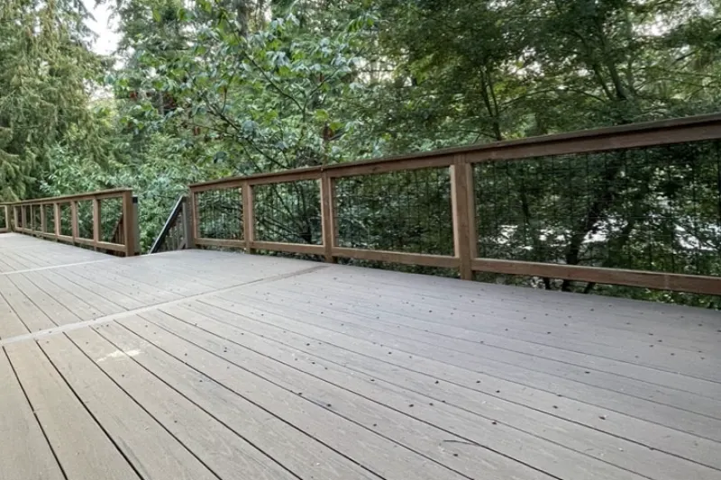 Composite deck with cedar and wire panel railings wrapping a corner beside tall evergreens, from a Mercer Island project by Volcano Builders