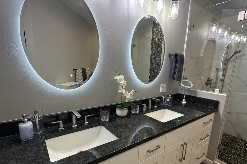 Double vanity with a dark granite top, two backlit oval mirrors and a glass shower, from a Kirkland bathroom remodel by Volcano Builders