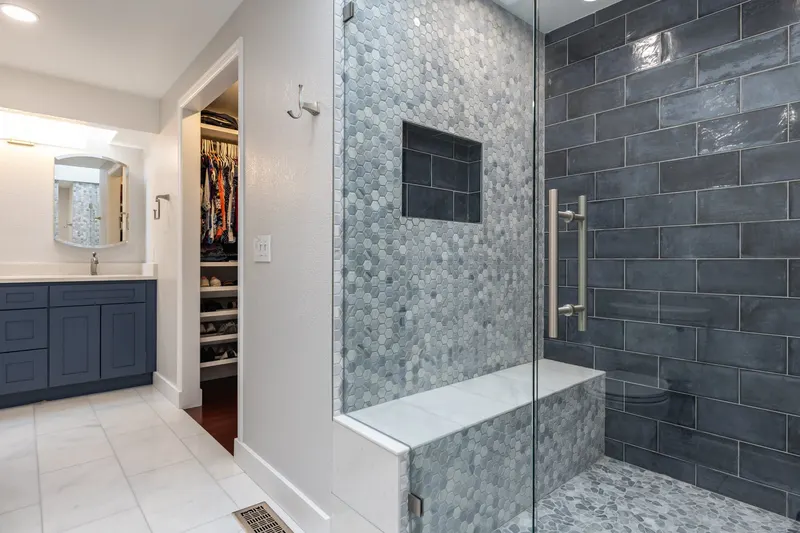 Walk-in shower with a glass panel, a tiled bench, hexagon and slate blue subway tile, and a navy vanity beyond, from a Redmond bathroom remodel by Volcano Builders