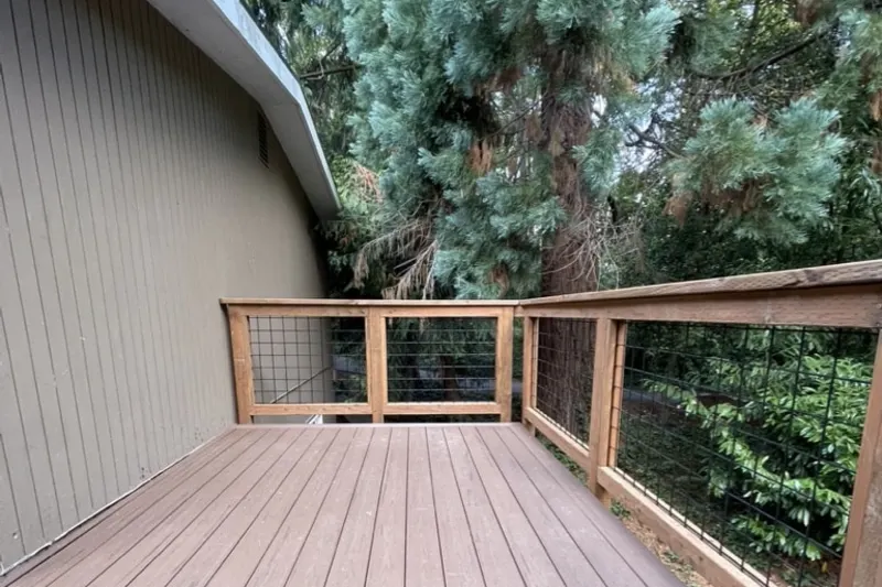 Composite side deck with a cedar rail and black wire panels beside grey siding and a blue spruce, from a Mercer Island deck remodel by Volcano Builders