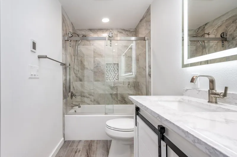 Bathroom with a marble look tiled tub surround, sliding glass door and white vanity, from a Tulalip Bay remodel by Volcano Builders