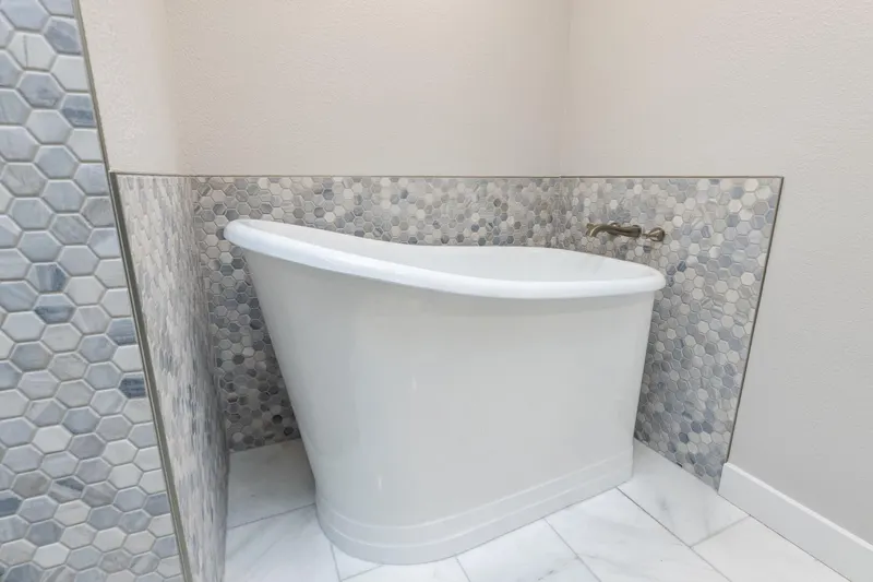 White soaking tub set in a tiled alcove of gray hexagon mosaic above a marble floor, a Redmond project by Volcano Builders