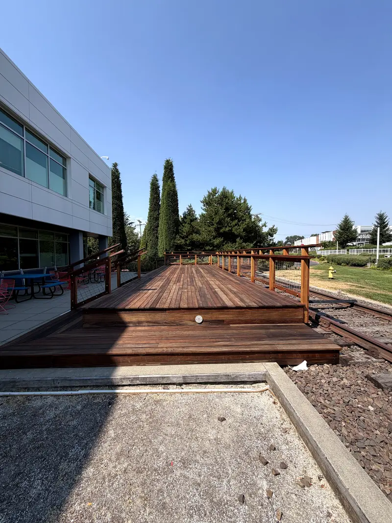 Refinished deck platform with wide steps and wood railing in front of a white office building under a clear sky