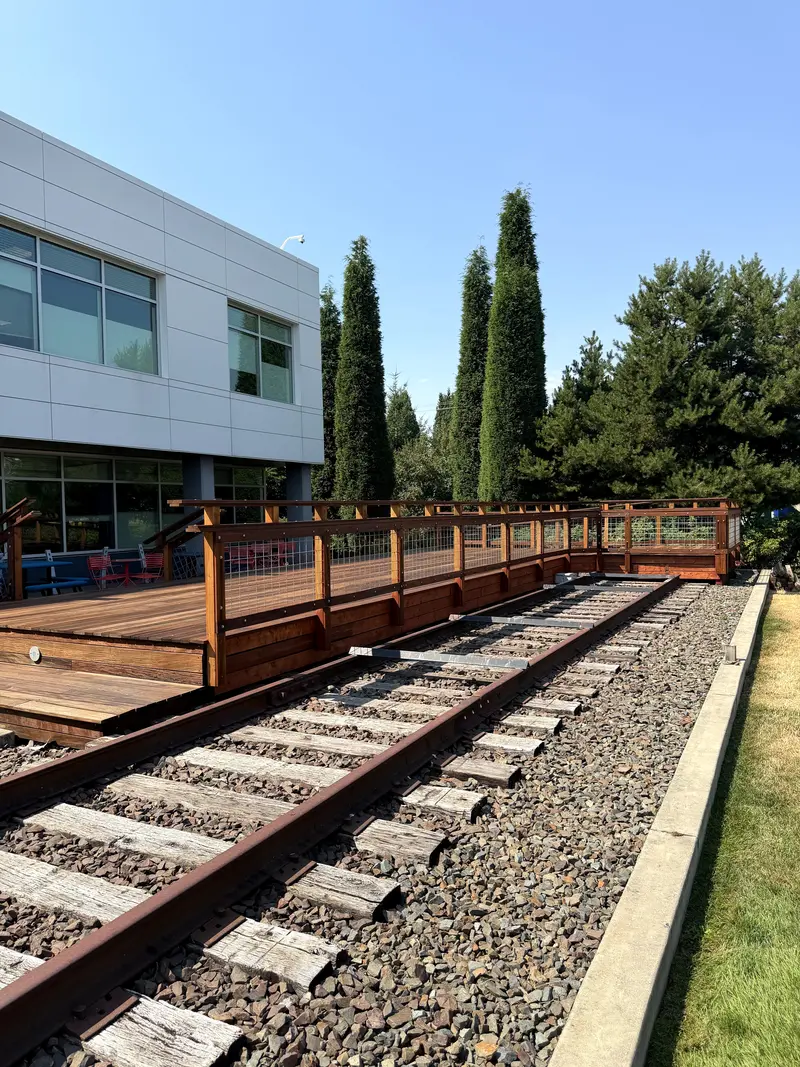 Refinished reddish brown hardwood deck with wood railing running beside old railroad track and gravel, next to an office building