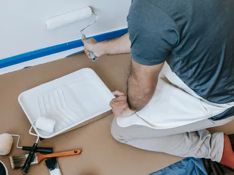 Worker rolling white paint onto a wall above blue painter's tape, with a paint tray, rollers and brushes on floor paper