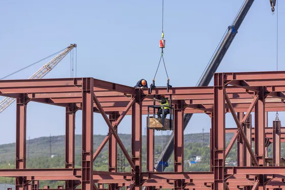 Two workers in hard hats fixing a red steel beam on a building frame, lifted by a crane