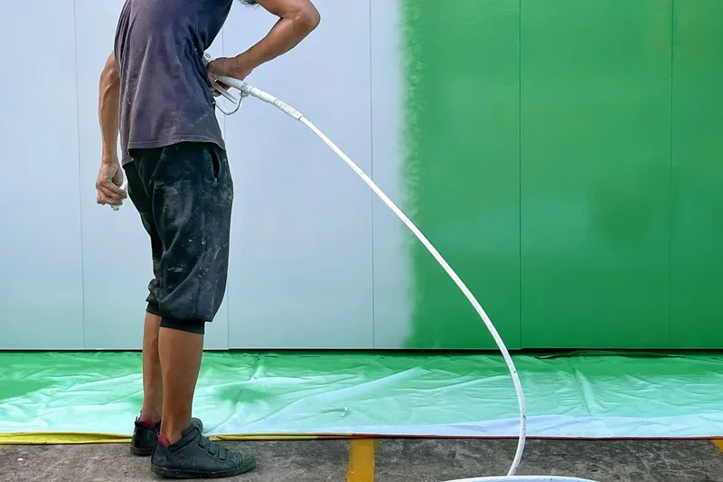 Painter holding an airless spray gun beside a metal panel wall that is changing from white to bright green, with a drop cloth spread over the pavement below