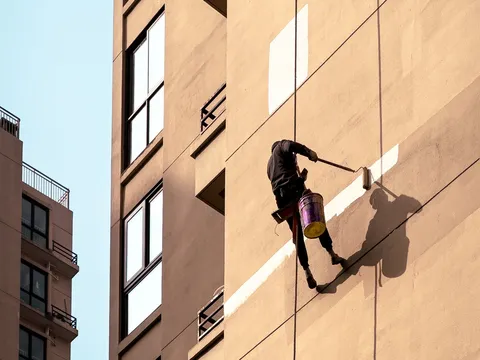 Painter hanging on ropes beside a tan multistory apartment facade, rolling a pale stripe of paint across the wall with a bucket at his hip