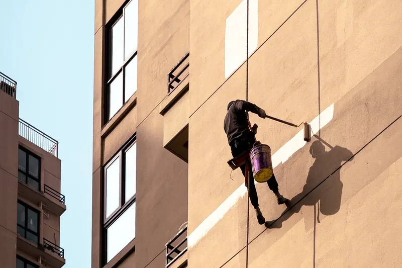 Painter hanging on ropes beside a tan multistory apartment facade, rolling a pale stripe of paint across the wall with a bucket at his hip
