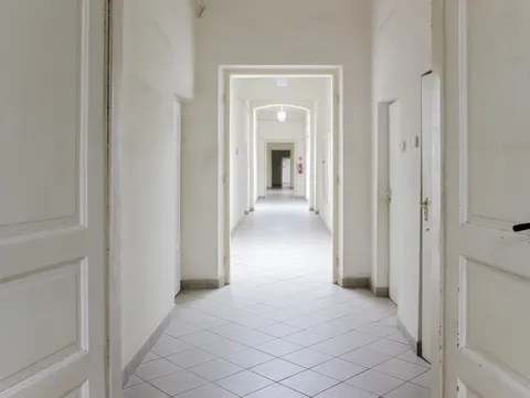Long white corridor seen through open paneled doors, with white walls, grey baseboards, a tiled floor and a row of doorways leading to the far end