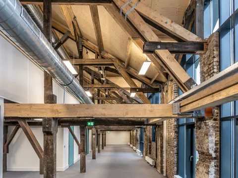 Renovated corridor in an older building with exposed timber trusses, brick piers, new white walls, carpet and a round steel air duct