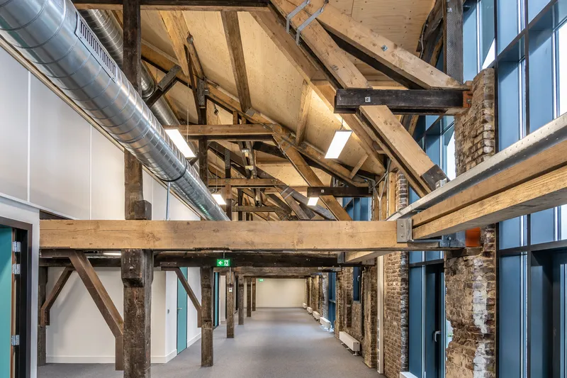 Renovated corridor in an older building with exposed timber trusses, brick piers, new white walls, carpet and a round steel air duct