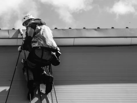 Black and white photo of a worker in a helmet and harness on a rope line at the edge of a metal roof