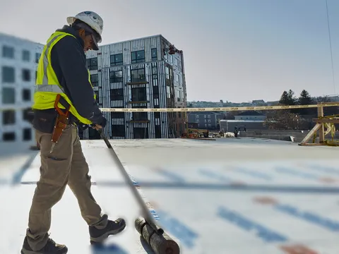 Roofer in a hard hat and safety vest pushing a weighted roller across a new membrane layer on a flat commercial roof