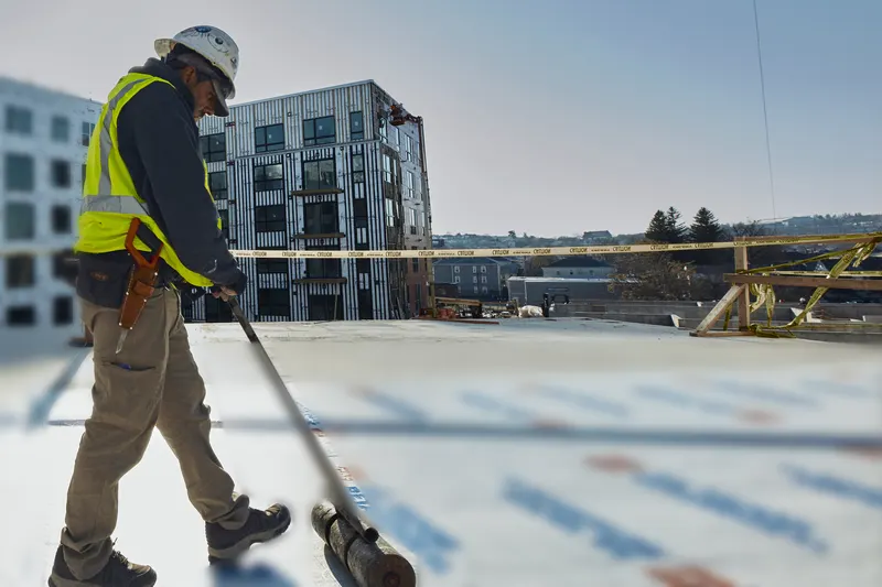 Roofer in a hard hat and safety vest pushing a weighted roller across a new membrane layer on a flat commercial roof