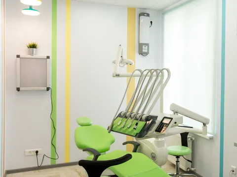 Dental treatment room with a green patient chair, delivery unit, overhead light and a window with a pale blind