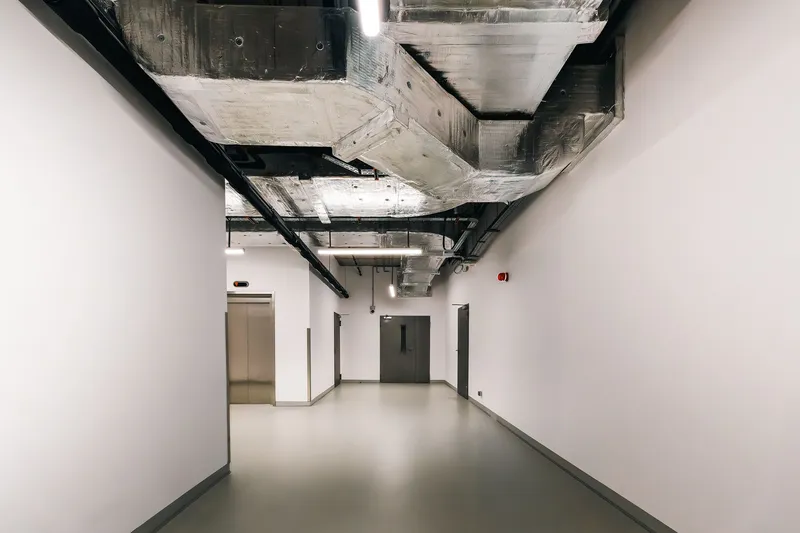 Commercial building corridor with insulated ductwork, cable trays and linear lights overhead, steel service doors and an elevator