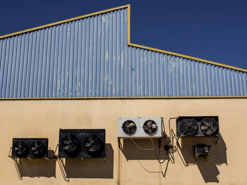 Row of condenser units with fans mounted on the stucco wall of an industrial building below corrugated metal cladding