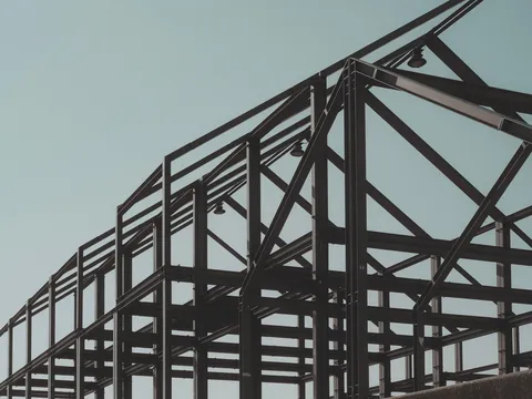Dark structural steel frame of a low building with columns, beams, diagonal bracing and a gabled roof line against a pale sky, no cladding yet