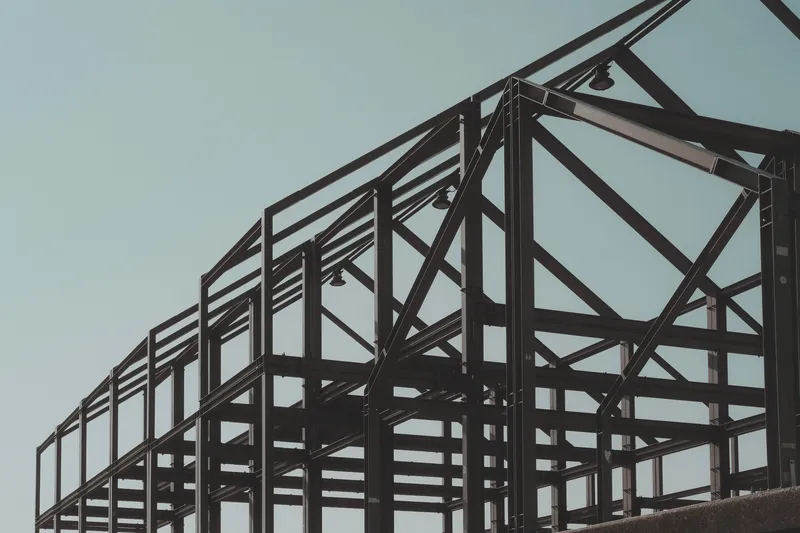 Dark structural steel frame of a low building with columns, beams, diagonal bracing and a gabled roof line against a pale sky, no cladding yet