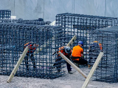Workers in orange shirts and hard hats tying tall rebar cages braced with timber props on a gravel site in front of a concrete wall
