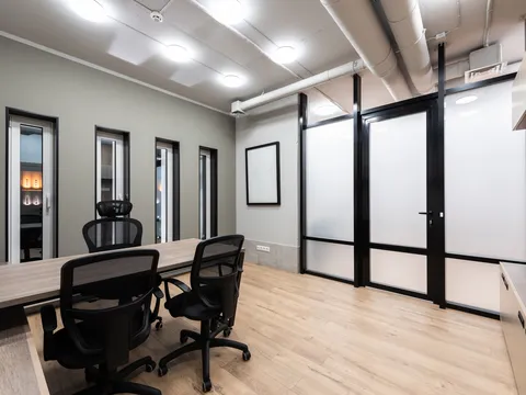 Small office meeting room with a wood table, black mesh chairs, light plank flooring, exposed white ductwork, round ceiling lights and a black framed frosted glass front with a door