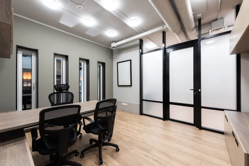 Small office meeting room with a wood table, black mesh chairs, light plank flooring, exposed white ductwork, round ceiling lights and a black framed frosted glass front with a door