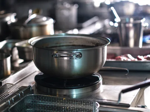 Stainless steel cook line with a large pot on a gas wok burner, fryer baskets in the foreground and stock pots and a sheet pan on the counters behind