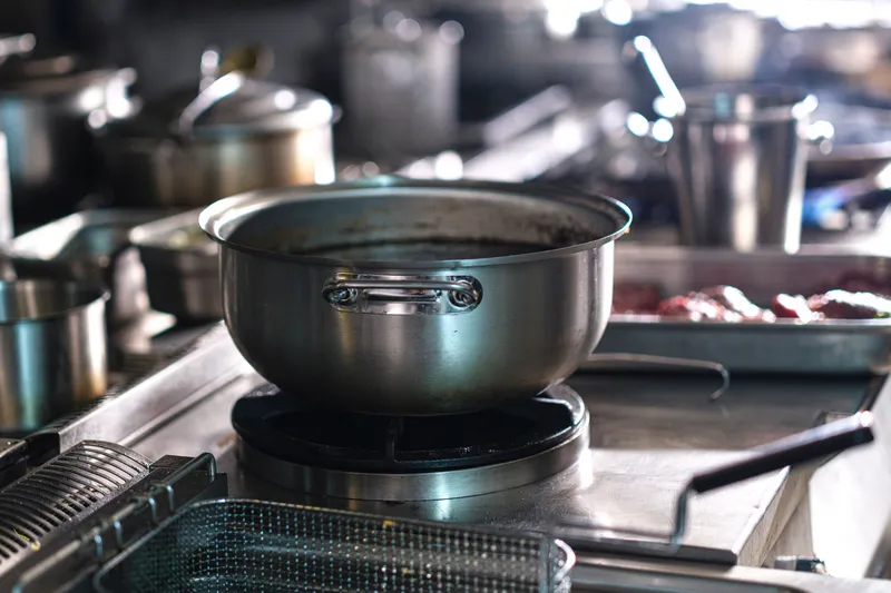Stainless steel cook line with a large pot on a gas wok burner, fryer baskets in the foreground and stock pots and a sheet pan on the counters behind
