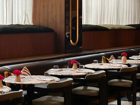 Empty restaurant dining room with round marble topped tables, upholstered chairs, a long leather banquette, small vases of red flowers and sheer curtains over wood paneled window seats