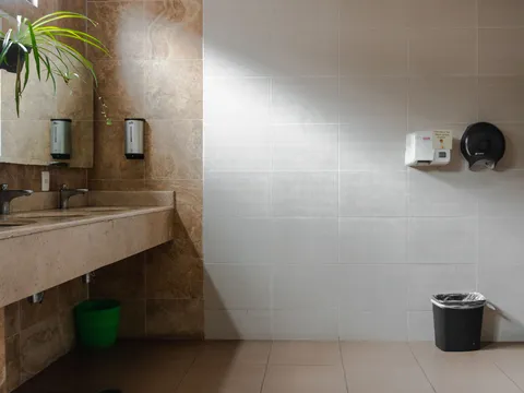 Commercial restroom with a stone counter holding two sinks, wall mounted soap dispensers, a hand dryer and a paper towel dispenser on a tiled wall