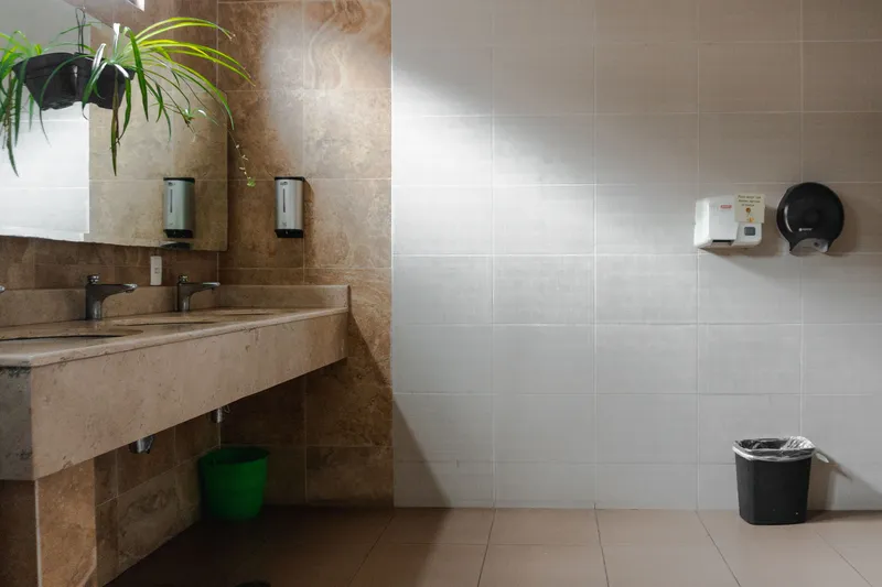 Commercial restroom with a stone counter holding two sinks, wall mounted soap dispensers, a hand dryer and a paper towel dispenser on a tiled wall