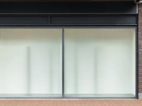 Vacant shopfront with two large whitened glass panes in dark metal frames, a plain dark fascia above and a brick pier at the right edge