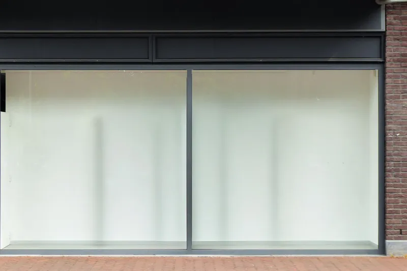 Vacant shopfront with two large whitened glass panes in dark metal frames, a plain dark fascia above and a brick pier at the right edge
