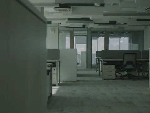 Empty open plan office with rows of desks and task chairs, low partitions, carpet tile, hung ceiling panels with cassette air units and glass walled rooms along the windows