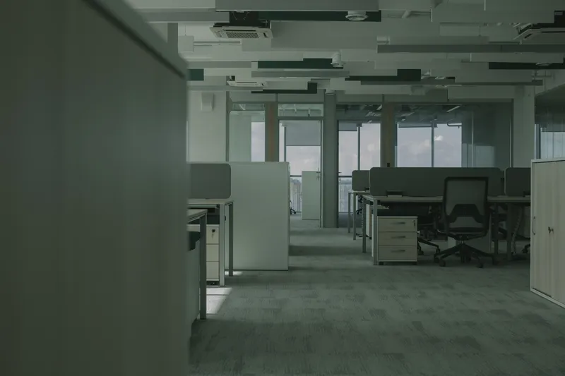 Empty open plan office with rows of desks and task chairs, low partitions, carpet tile, hung ceiling panels with cassette air units and glass walled rooms along the windows